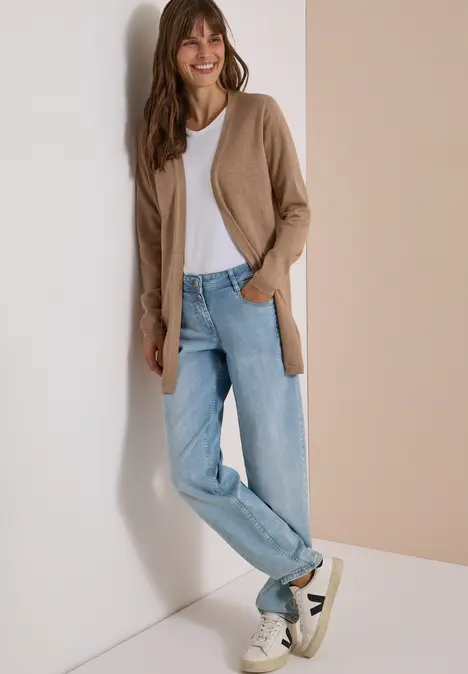 Eine Frau lehnt an einer Wand, trägt hellblaue Jeans, weißes T-Shirt, braunen Cardigan und weiße Sneakers.