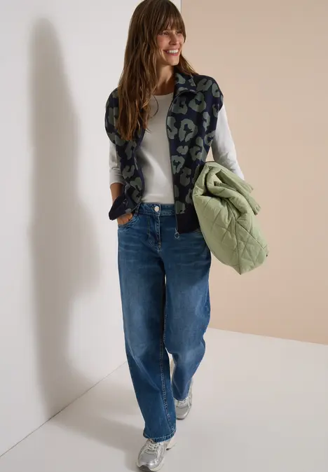 Fröhliche Frau in Jeans, weißem Langarmshirt und gemusterter Weste, hält eine hellgrüne Steppjacke.
