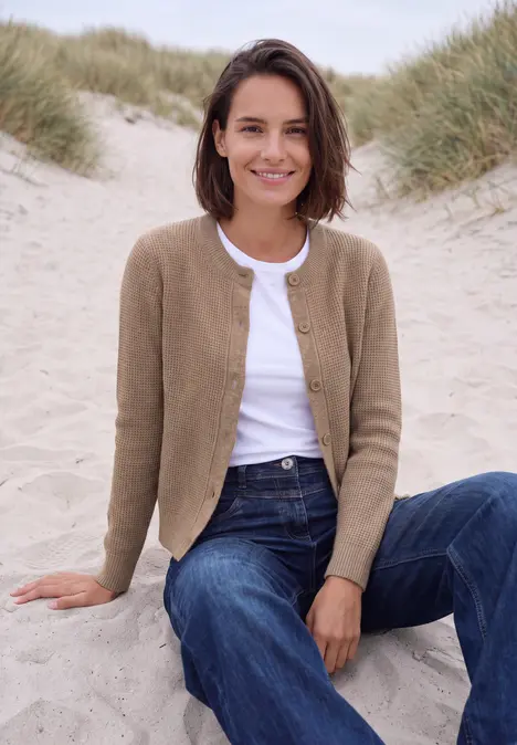 Eine lächelnde Frau in Jeans und Cardigan sitzt entspannt auf Dünen im Strandhintergrund.
