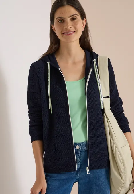 Lächelnde Frau in dunkelblauer Kapuzenjacke, mintgrünem Top und Jeans mit beiger Umhängetasche.