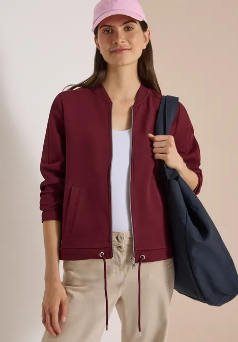Frau in burgundfarbener Jacke, weißem Top, beiger Hose, rosa Mütze und großer blauer Tasche.