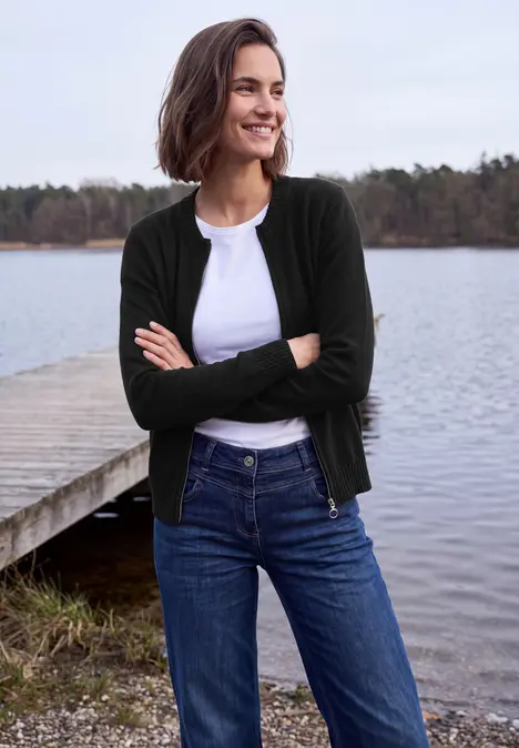 Frau in Jeans, weißem Shirt und dunklem Cardigan steht lächelnd mit verschränkten Armen am Seeufer.
