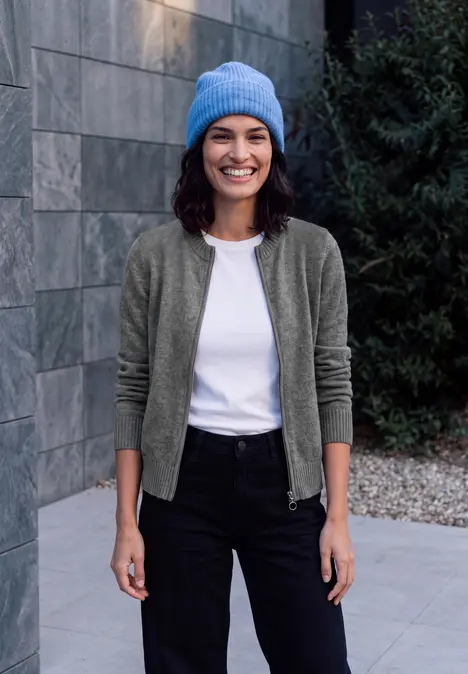 Eine lächelnde Frau in blauer Mütze, weißem Shirt, grüner Strickjacke und schwarzer Hose vor einer Steinwand.