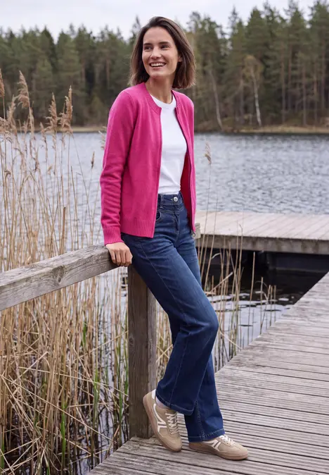 Eine lächelnde Frau in pinkem Cardigan, weißem Shirt und Jeans steht auf einem Steg am See und lehnt am Geländer.