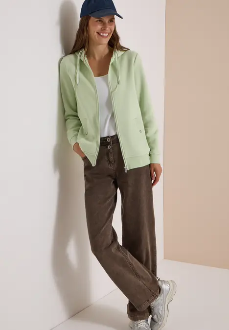 Junge Frau lehnt an Wand, trägt grünen Cardigan, weißes Top, braune Cordhose, weiße Sneaker und blaue Mütze.