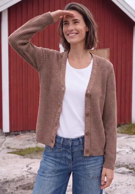 Eine lächelnde Frau in Cardigan und Jeans steht vor einem roten Holzhaus im Freien.