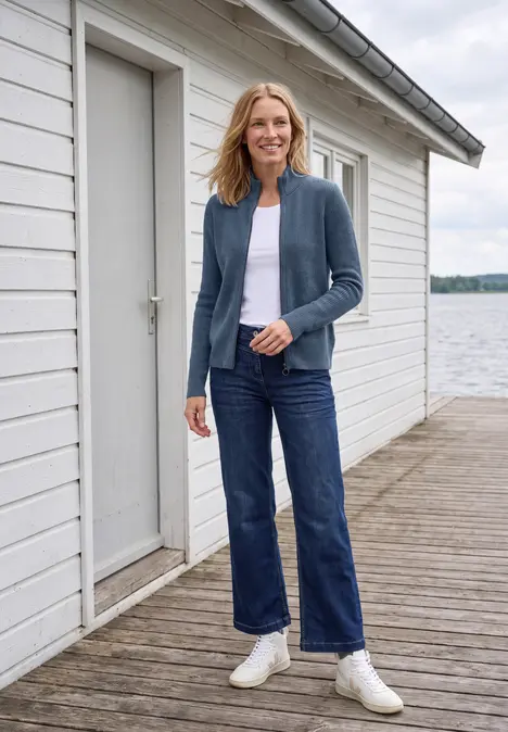 Frau in Jeans, weißem Shirt und Cardigan steht lächelnd auf einem Steg vor einem weißen Bootshaus am See.