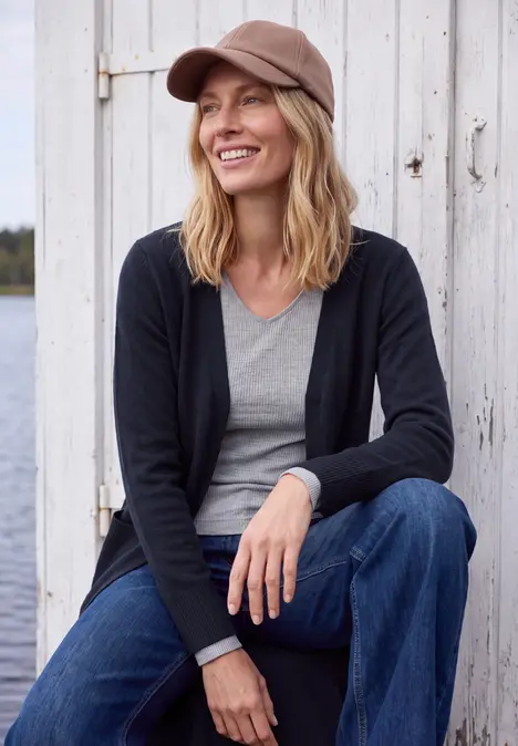 Eine lächelnde Frau mit Schirmmütze, grauem Shirt, schwarzer Jacke und Jeans sitzt vor einer Holzwand am Wasser.