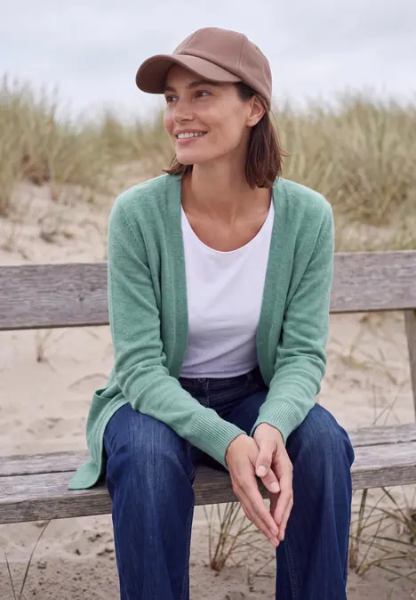 Eine Frau mit Mütze und grünem Cardigan sitzt lächelnd auf einer Bank vor Dünen am Strand.