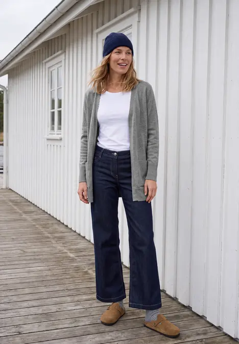 Frau in lässigem Outfit (Beanie, Cardigan, weißes T-Shirt, weite Jeans, Loafers) vor weißem Holzhaus.