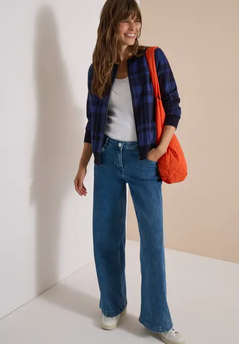 Frau in kariertem Hemd, weißem Shirt, Jeans, weißen Sneakern und orangefarbener Tasche vor neutralem Hintergrund.