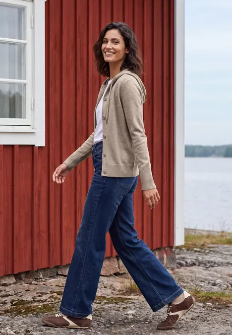 Frau in Jeans und Jacke spaziert fröhlich an einem roten Holzhaus am Wasser.