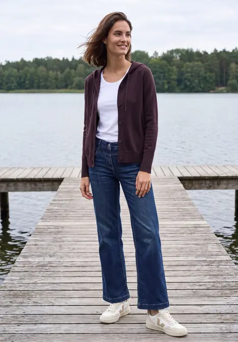 Frau in weißem T-Shirt, lila Hoodie und Jeans steht lächelnd auf einem Steg am See.