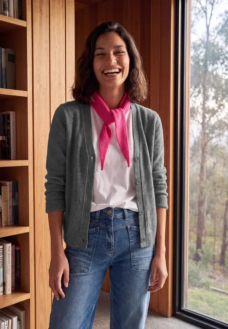 Eine lächelnde Frau in Jeans, T-Shirt und grauer Strickjacke steht locker vor einem Fenster im Holzinterieur.
