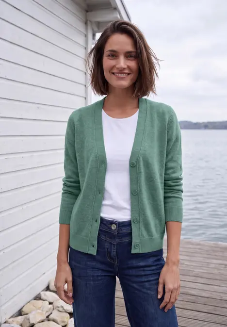 Lächelnde Frau im grünen Cardigan und Jeans lehnt draußen an einer Steinwand.
