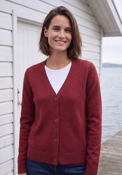 Eine Frau in rotem Cardigan und weißem Shirt lächelt auf einem Steg vor einem weißen Haus am Wasser.