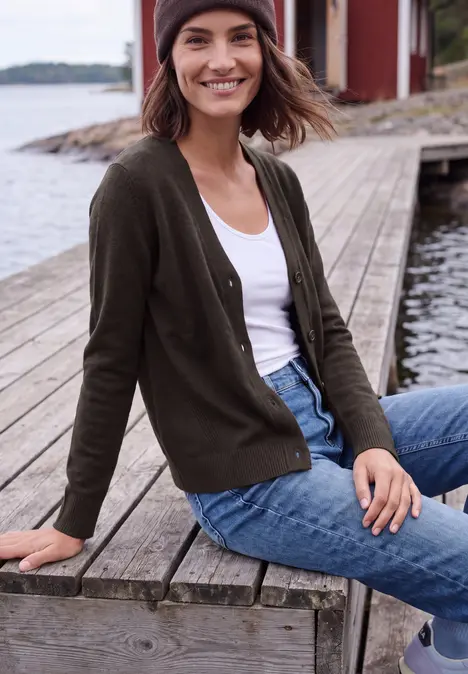 Junge Frau sitzt lächelnd auf einem Holzsteg am Wasser in Strickjacke, Jeans und Mütze.