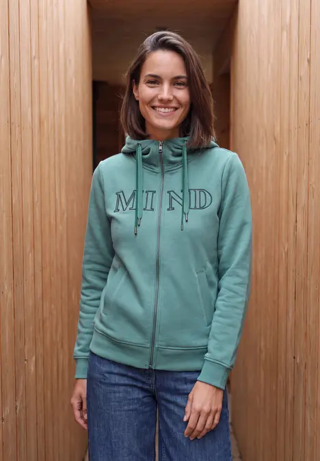 Eine lächelnde Frau in grünem Kapuzenpullover mit 'MIND'-Schriftzug vor hölzerner Wand.