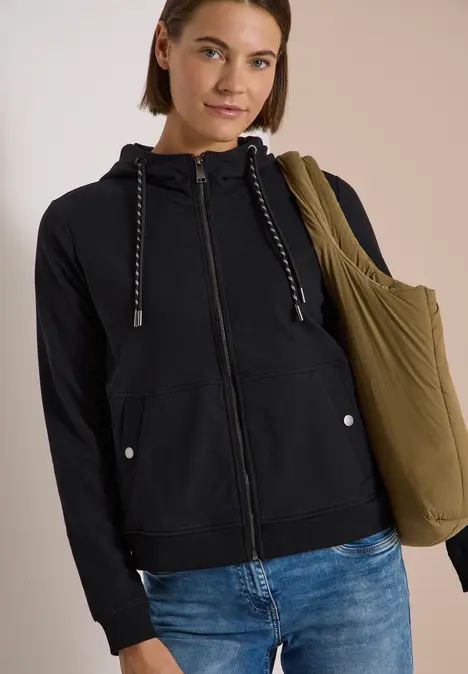 Frau in Jeans und schwarzem Hoodie trägt olivgrüne Tasche vor hellem Hintergrund.