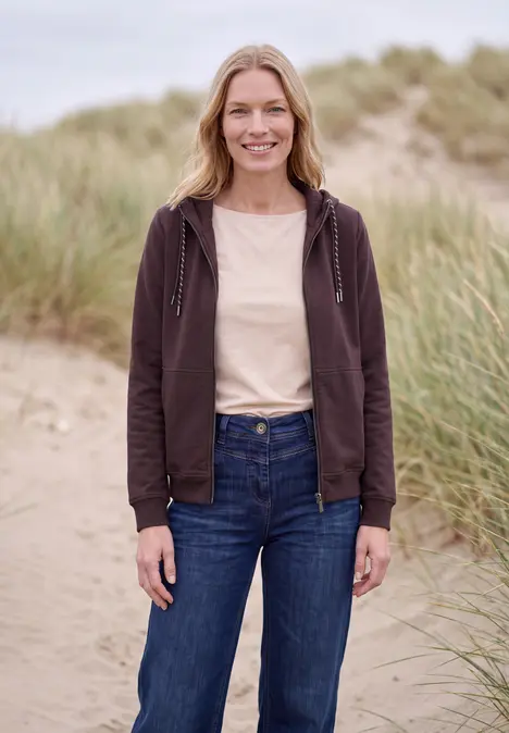 Eine lächelnde Frau in brauner Kapuzenjacke und Jeans steht auf Dünen mit Strandgras im Hintergrund.