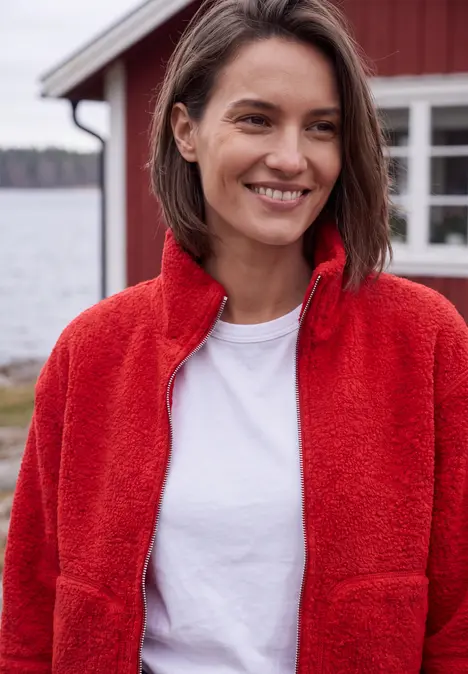 Eine lächelnde Frau in roter Fleecejacke und weißem T-Shirt steht vor einem roten Haus am See.
