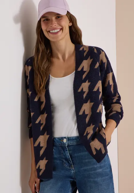 Lächelnde Frau in Hahnentritt-Strickjacke, weißem Top, Jeans und Cap
