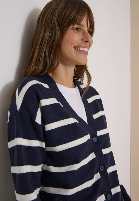 Frau lächelt und trägt einen marineblauen Cardigan mit weißen Streifen und ein weißes Shirt.
