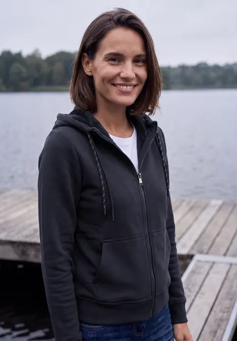 Eine junge Frau in Kapuzenjacke steht auf einem Steg am See und lächelt in die Kamera.