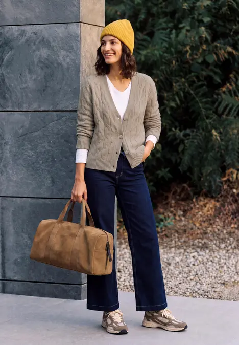 Frau in gelber Mütze, grauem Cardigan und blauer Hose mit Reisetasche vor modernem Gebäude und Bäumen.