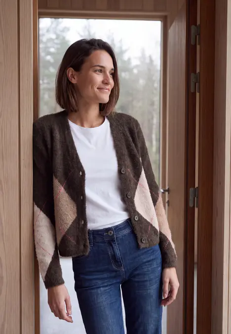Eine Frau in Jeans, weißem Shirt und kariertem Cardigan lehnt an einem Türrahmen und blickt nach draußen.