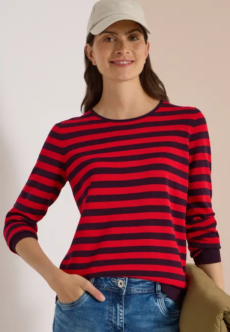 Frau mit rotem-schwarzem Ringelshirt, Jeans und Baseballcap, lächelnd, eine Hand in der Tasche.