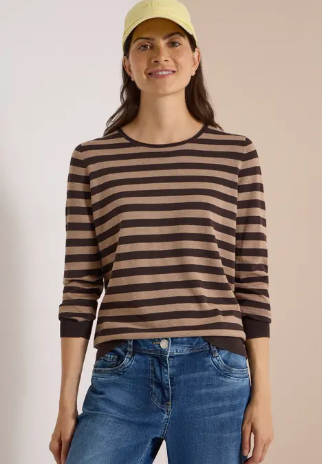 Eine lächelnde Frau trägt einen braun-schwarz gestreiften Pullover, Jeans und Baseballcap.