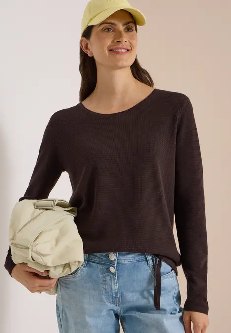 Junge Frau mit Cap, dunkelbraunem Pullover, Jeans und beiger Umhängetasche lächelt.