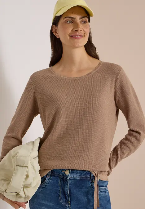 Frau mit hellbraunem Pullover und Cap, lässig mit Tasche über dem Arm, lächelnd zur Seite blickend.