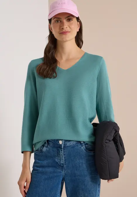 Frau mit rosa Cap, grünem V-Ausschnitt-Pullover, blauen Jeans und schwarzer Jacke in der Hand.