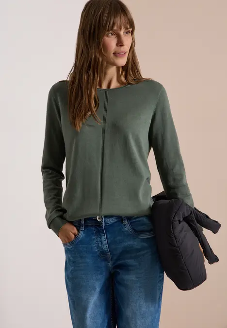 Frau in grünem Pullover, Jeans und hält eine dunkle Jacke in der Hand.