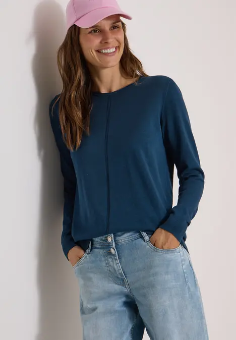 Eine Frau mit blauem Langarmshirt, Jeans und rosa Cap lehnt an einer Wand und lächelt.