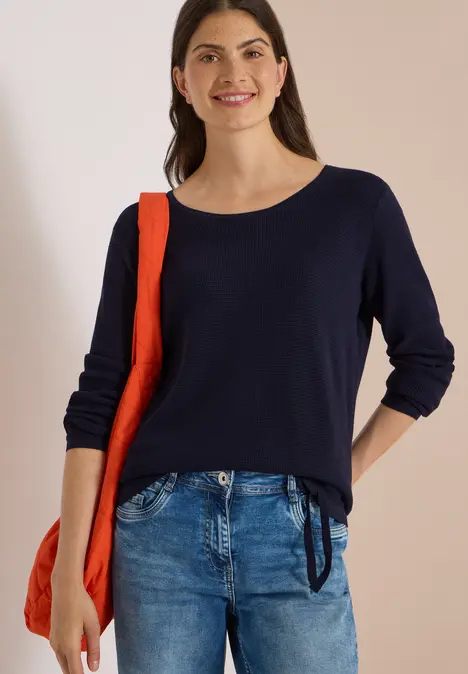 Junge Frau in dunkelblauem Pullover und Jeans trägt eine rote Umhängetasche und lächelt.