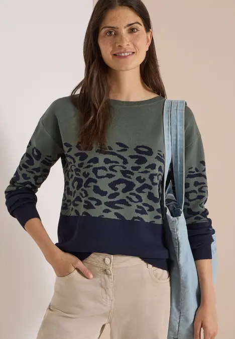 Frau mit grün-blauem Pullover mit Leopardenmuster, heller Hose und Jeans-Tasche lächelt in die Kamera.