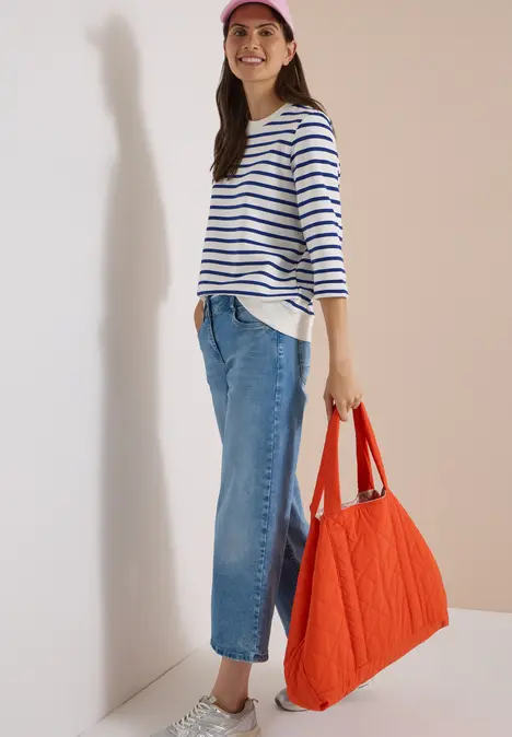 Frau in Streifenshirt, Jeans und roter Tasche lehnt an weißer Wand und lächelt.