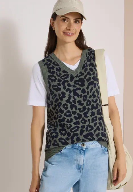Frau in Schirmmütze trägt leo-print Weste über weißem Shirt und Jeans, mit Schultertasche, lächelt.