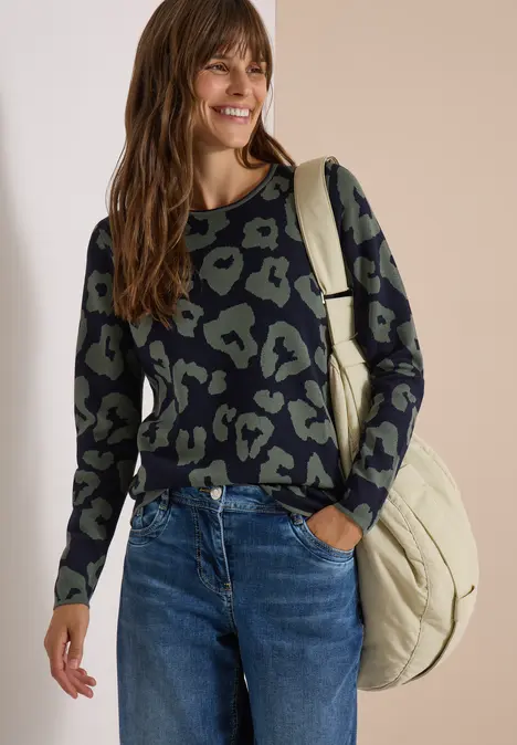 Frau mit gemustertem Pullover in Grün und Schwarz, blauer Jeans und Beuteltasche, lächelnd vor beigem Hintergrund.