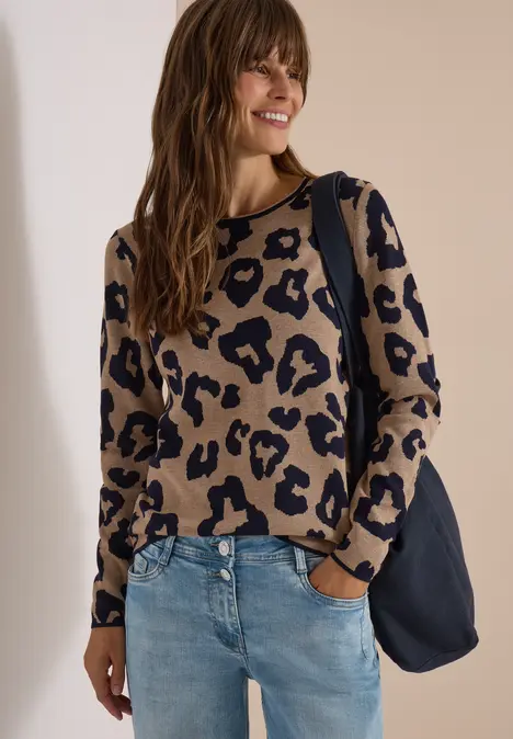 Frau mit beige-schwarzem Leo-Pullover, blauer Jeans und dunkelblauer Tasche lächelt.