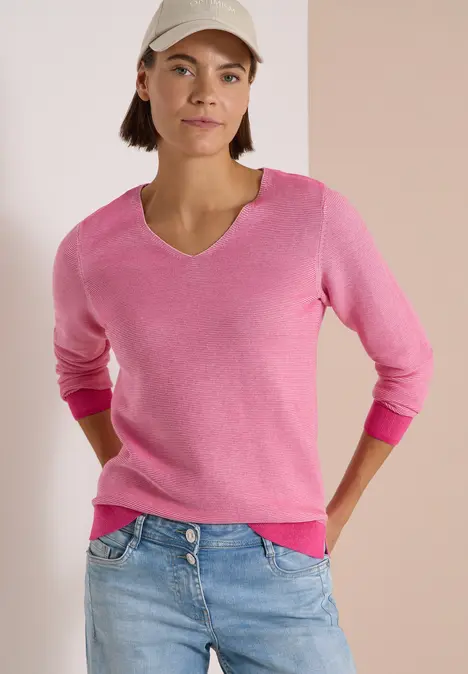 Vrouw met roze V-hals trui, lichte spijkerbroek en pet voor neutrale achtergrond.