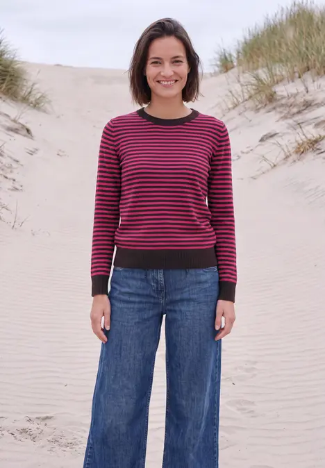 Eine Frau steht in Jeans und gestreiftem Pullover auf einer Düne am Meer.