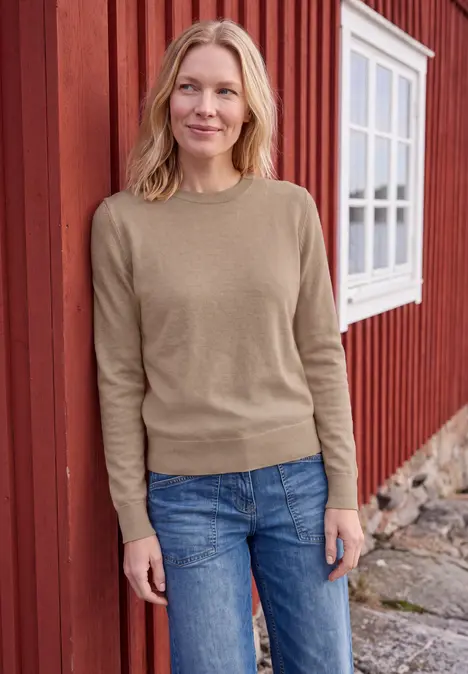 Frau in beigem Pullover und Jeans steht vor rotem Haus mit Fenster.