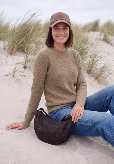 Junge Frau sitzt am Strand auf Sanddüne, trägt Mütze, Pullover, Jeans und Tasche, dahinter Dünengras.