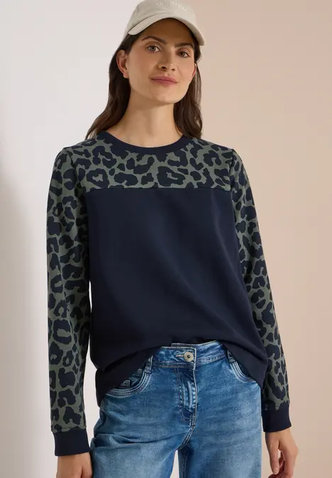 Frau in dunkelblauem Pullover mit Leopardenmuster an Ärmeln und Jeans, mit Basecap vor neutralem Hintergrund.