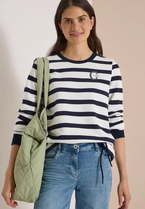 Frau mit dunkelblau-weiß gestreiftem Pullover, Jeans und grüner gesteppter Umhängetasche.