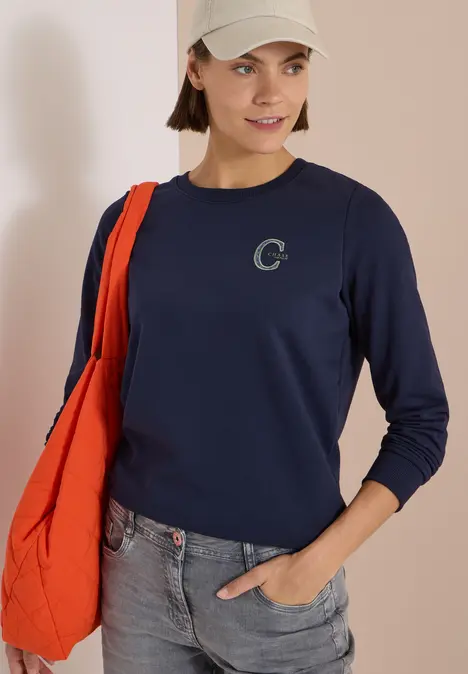 Frau mit beigem Cap, dunkelblauem Sweatshirt mit kleinem C-Logo, Jeans und oranger Tasche vor schlichtem Hintergrund.